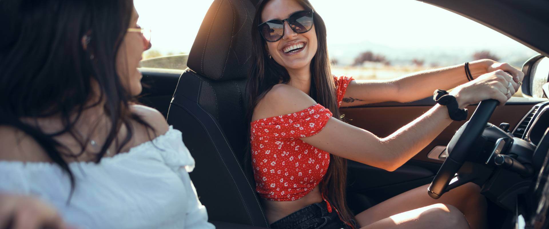 A smiling woman wearing sunglasses is driving a car, with a passenger raising her arms joyfully in the background, all against a sunny outdoor backdrop.